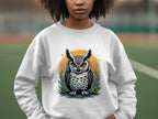 A person sports a Garment Graphics white sweatshirt featuring a striking owl design with vivid eyes and intricate feathers. The scene is set on a sports field, and the unisex sizing guarantees an ideal fit for everyone.
