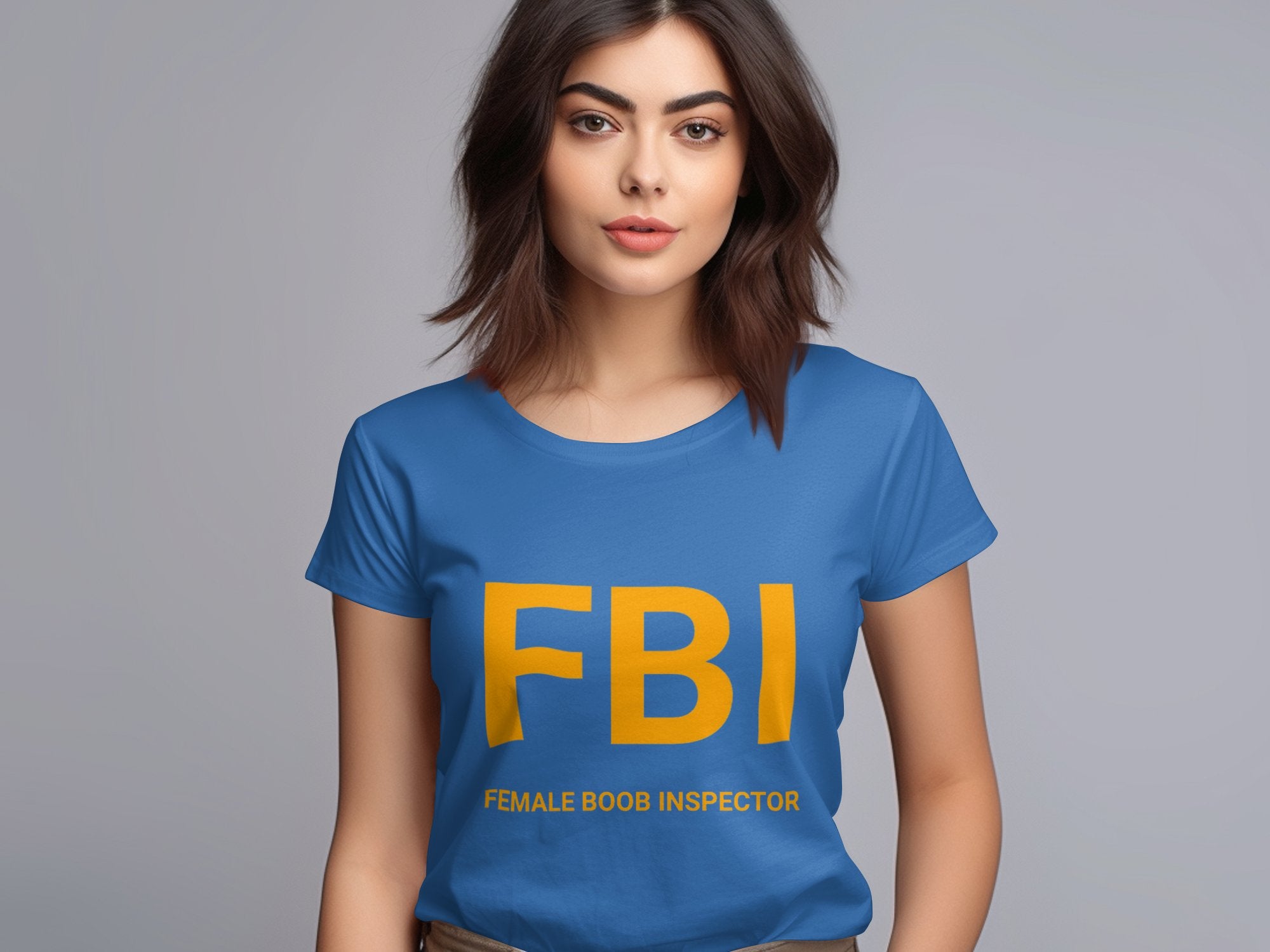 A woman with shoulder-length brown hair wears a Garment Graphics blue T-shirt featuring large yellow letters FBI and Female Boob Inspector. Against a plain gray backdrop, this playful womens tee makes an amusing gag gift sure to start conversations.