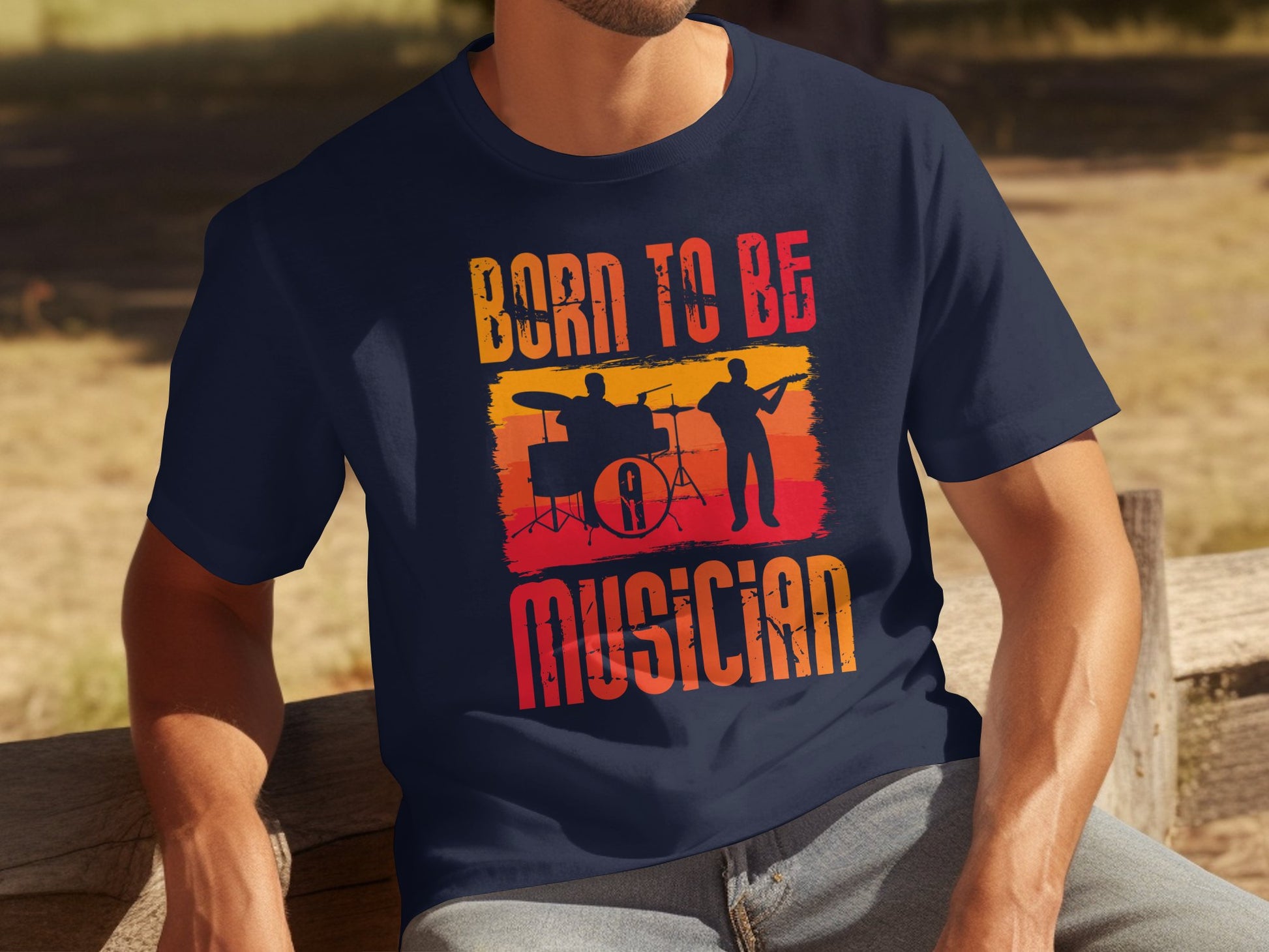 A person sits on a bench wearing Garment Graphics iconic Born To Be Musician T-shirt. This navy blue shirt, adored by music lovers, features silhouettes of a drummer and guitarist against a sunset gradient with bold letters celebrating the love for rhythm and melody.