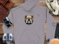 A gray hoodie from Garment Graphics, showcasing a bold bulldog with a spiked collar, is neatly placed on a wooden surface. Its surrounded by items such as a camera, binoculars, leather book, watch, gloves, and climbing carabiner.