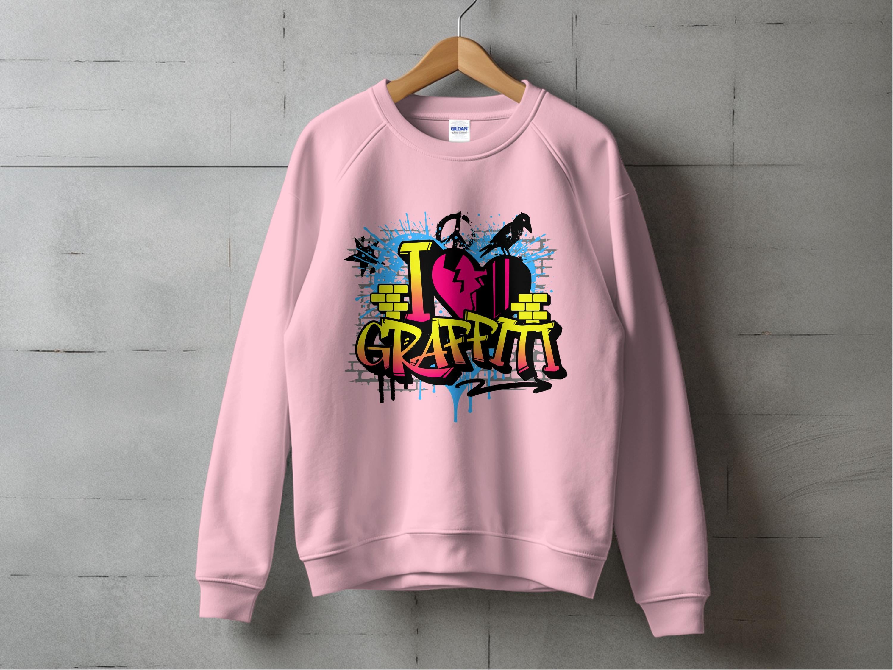 A pink sweatshirt by Garment Graphics hangs on a wooden hanger against a concrete wall, featuring street art flair with graffiti-style elements, bold I Love Graffiti text, and artistic brick and paint splatter touches.
