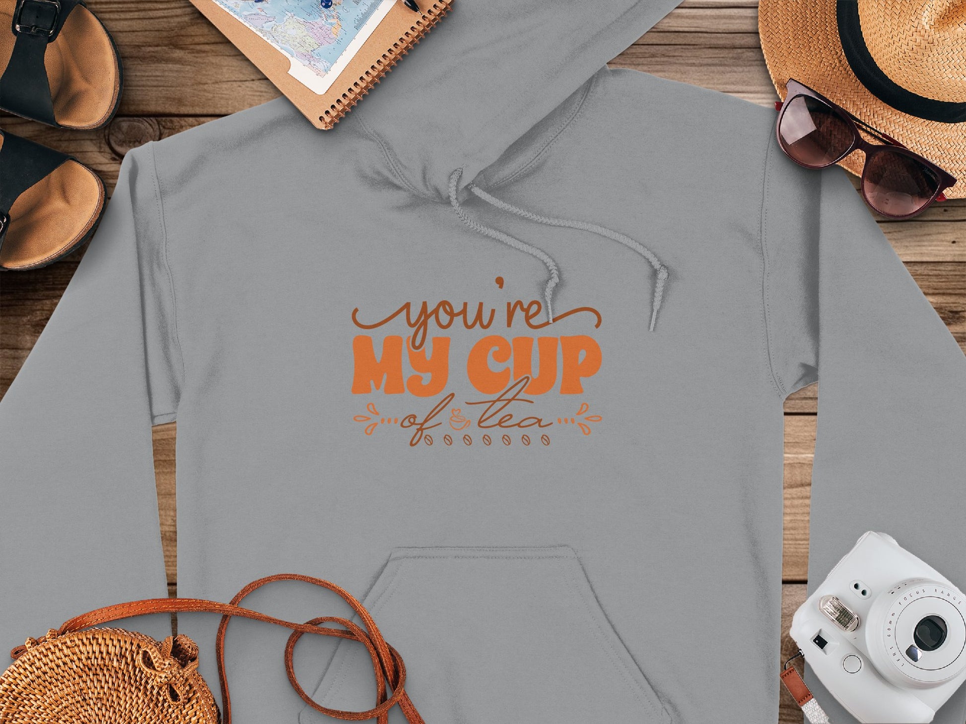 A cozy gray hoodie from Garment Graphics with Youre my cup of tea in orange, styled among a camera, straw hat, sandals, woven bag, and a spiral notebook with a map, ideal for capturing lifes cozy moments.