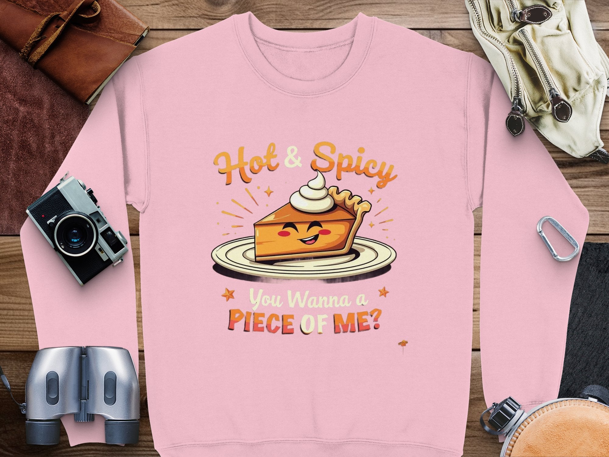 A pink unisex sweatshirt from Garment Graphics features a cartoon pie slice with whipped cream, reading Hot & Spicy. You Wanna a Piece of Me? It is arranged alongside a camera, binoculars, and other items.