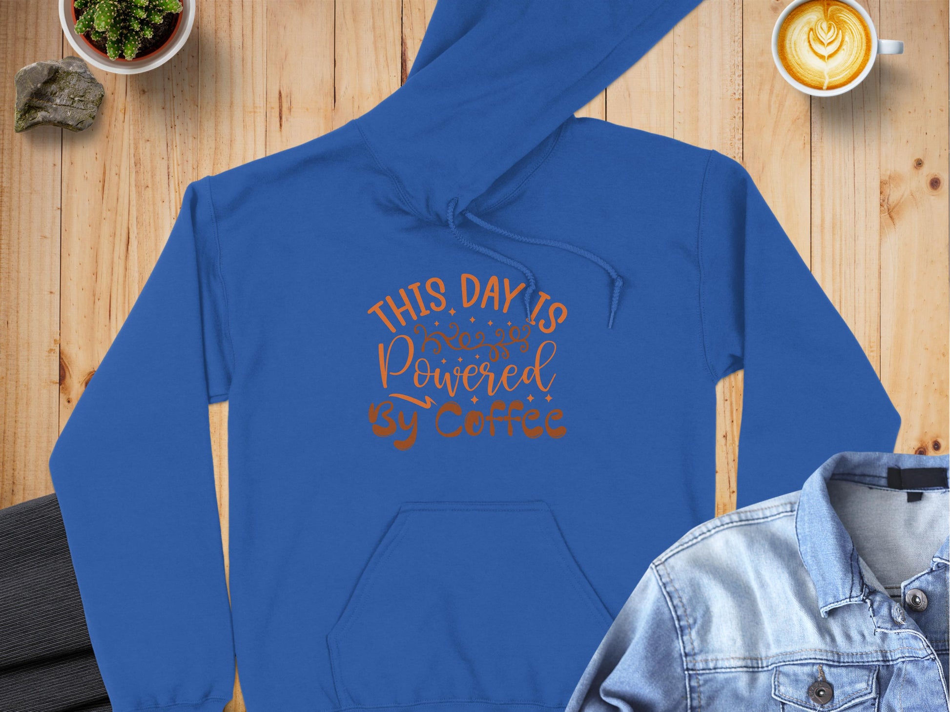 Garment Graphics blue hoodie featuring This Day is Powered by Coffee rests on wood. A denim jacket adds style on the right, and a latte art cup sits in the top corner, completing this perfect scene for coffee lovers.