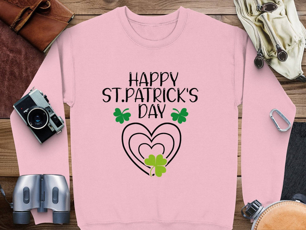 A festive pink sweatshirt from Garment Graphics, featuring Happy St. Patricks Day, is adorned with vivid green shamrocks and hearts. It is surrounded by a camera, binoculars, leather pouch, and gloves on a wooden surface.