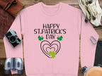 A festive pink sweatshirt from Garment Graphics, featuring Happy St. Patricks Day, is adorned with vivid green shamrocks and hearts. It is surrounded by a camera, binoculars, leather pouch, and gloves on a wooden surface.