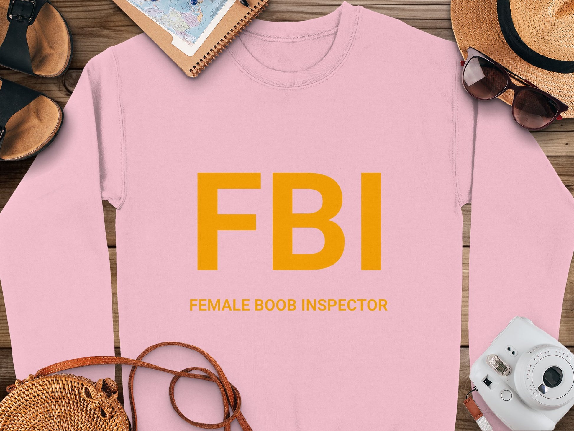 On a wooden surface, this Garment Graphics sweatshirt highlights bold yellow FBI letters with FEMALE BOOB INSPECTOR below. Surrounded by a hat, sunglasses, camera, sandals, and notebook, it offers unisex sizing for the daring wearer.