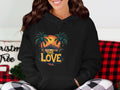 A person expresses their unique style by wearing a Garment Graphics black graphic hoodie featuring a vibrant tropical sunset with two palm trees and the phrase One Love, sitting on a couch with a Christmas Tree pillow and plaid pants.