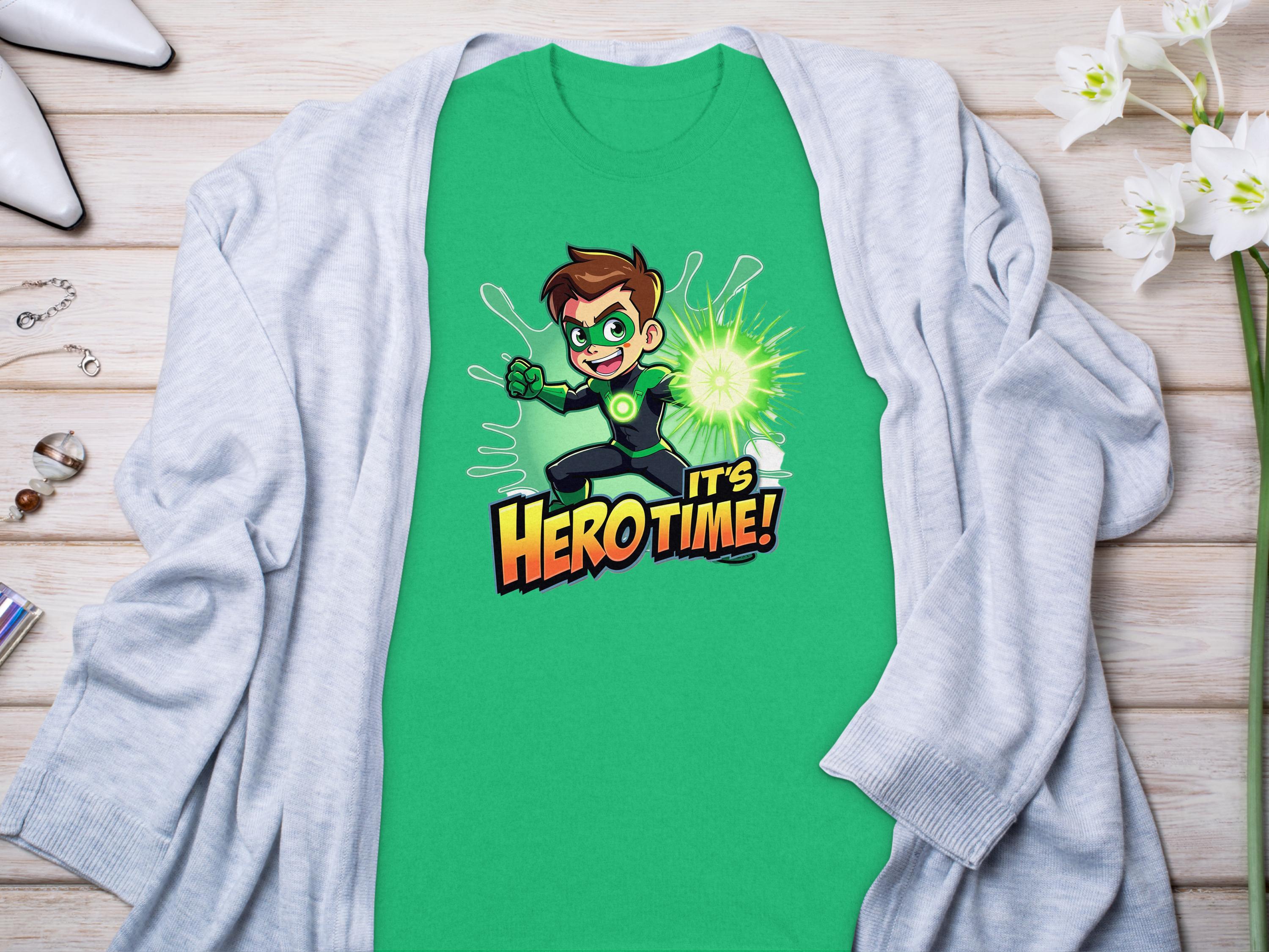 A vibrant green t-shirt from Garment Graphics showcases a superhero cartoon character holding a glowing green energy ball with the bold text Its Hero Time! Its paired with a light gray cardigan amidst jewelry and flowers on a wooden surface.