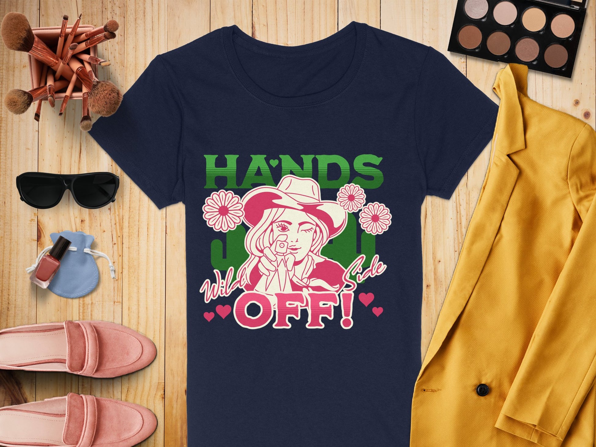 A navy Garment Graphics T-shirt with Hands Off! and a cowboy in a hat, surrounded by flowers, is styled on wood with a yellow jacket, makeup palette, brushes, sunglasses, purse, nail polish, and pink shoes.