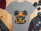 The Garment Graphics gray T-shirt, ideal for a beach day, features a tropical sunset with palm trees, bird, and One Love text. It’s placed on wood alongside winter wear like a red knit hat, gloves, and plaid shirt.