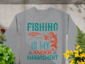 Garment Graphics presents a gray sweatshirt with the bold message Fishing is my anger management in vibrant colors. Ideal for fishing enthusiasts, it features a fish and hook illustration among greenery, accented with small yellow and red flowers.