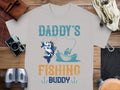 Ideal for fishing enthusiasts, this Garment Graphics gray T-shirt features Daddys Fishing Buddy in bold blue and orange. The design depicts a man fishing from a boat with a hooked fish, photographed on wood surrounded by a camera, binoculars, and leather items.