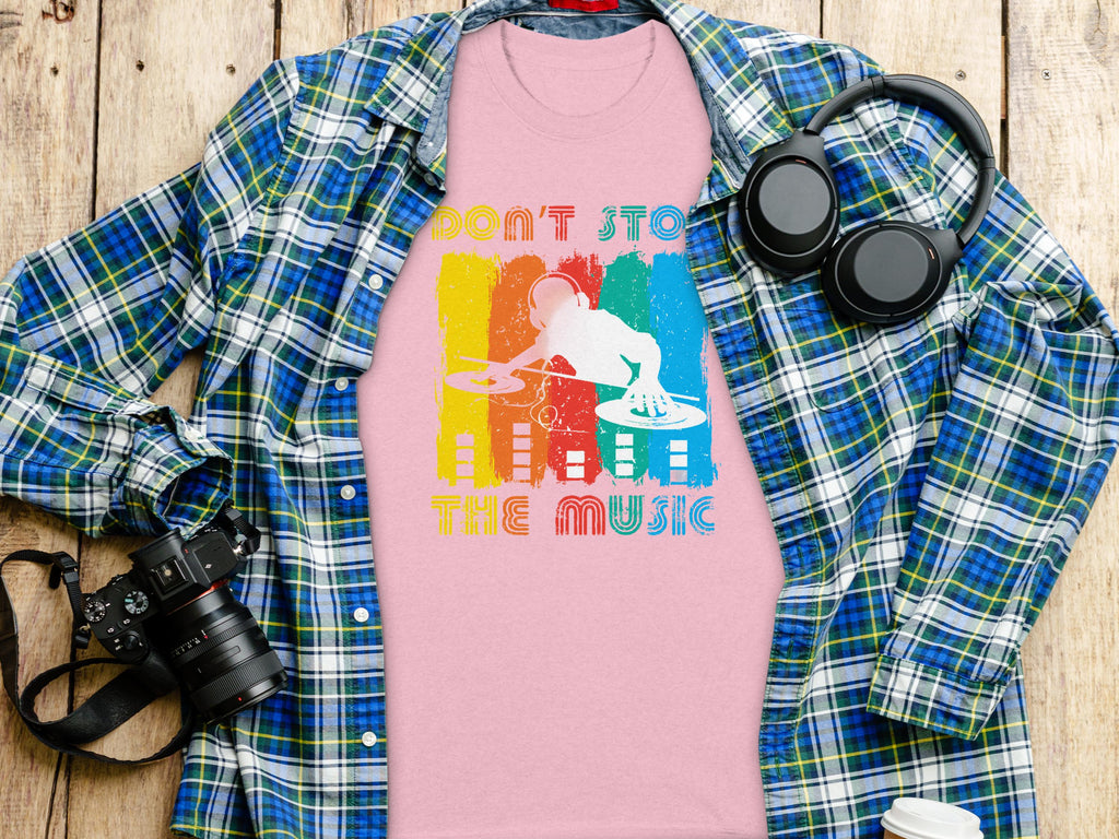 The Garment Graphics pink t-shirt with a DJ graphic and Dont Stop the Music text is perfect for music lovers. Its laid under a blue plaid shirt, surrounded by headphones, a camera, and a travel mug on a wooden surface.