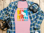 The Garment Graphics pink t-shirt with a DJ graphic and Dont Stop the Music text is perfect for music lovers. Its laid under a blue plaid shirt, surrounded by headphones, a camera, and a travel mug on a wooden surface.