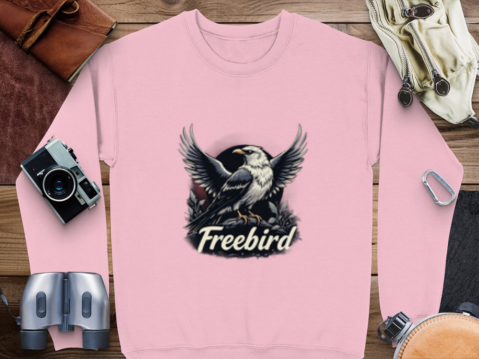 A pink sweatshirt from Garment Graphics features an eagle with outstretched wings and Freebird text. Its laid on wood with a camera, binoculars, a bag, and more—an ideal unisex Freebird Graphic Sweatshirt for adventurers.