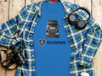 A blue t-shirt from Garment Graphics with a Scania truck design sits on wood. Its layered over an open plaid shirt, accompanied by headphones, a camera, and a coffee cup—ideal for truck enthusiasts.