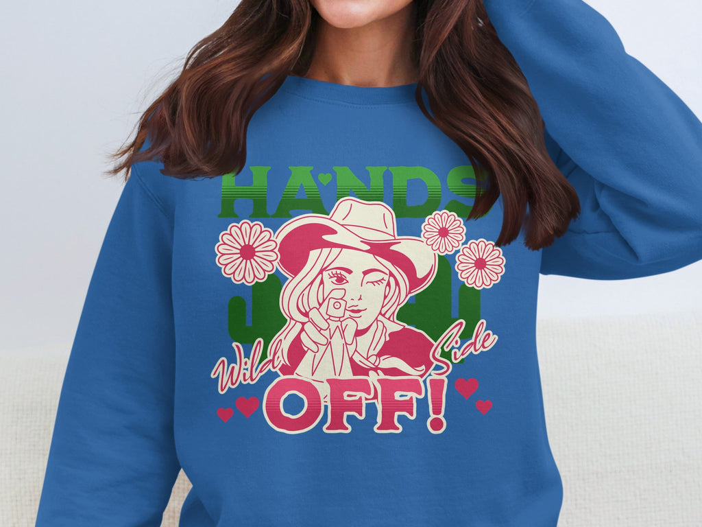 A woman sports a blue, loose-fit sweatshirt from Garment Graphics featuring a unique design: a woman in a hat, pink flowers, green HANDS, and red Wild Side OFF! text with red hearts. Ideal for standout style from the Hands off Wild Side Sweatshirts collection.