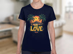 A person is wearing a Garment Graphics black T-shirt with a vibrant design of two palm trees, a tropical sunset behind a mountain, and a soaring bird. The words One Love are in bold yellow on the front, all set against an indoor background that evokes beach outing vibes.
