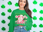 A woman beams in her loose-fit green Garment Graphics sweatshirt, featuring a hat-wearing character surrounded by flowers, with the text Hands Off Wild Side Sweatshirts. She stands against a shamrock-decorated wall, stylishly pairing it with denim shorts.