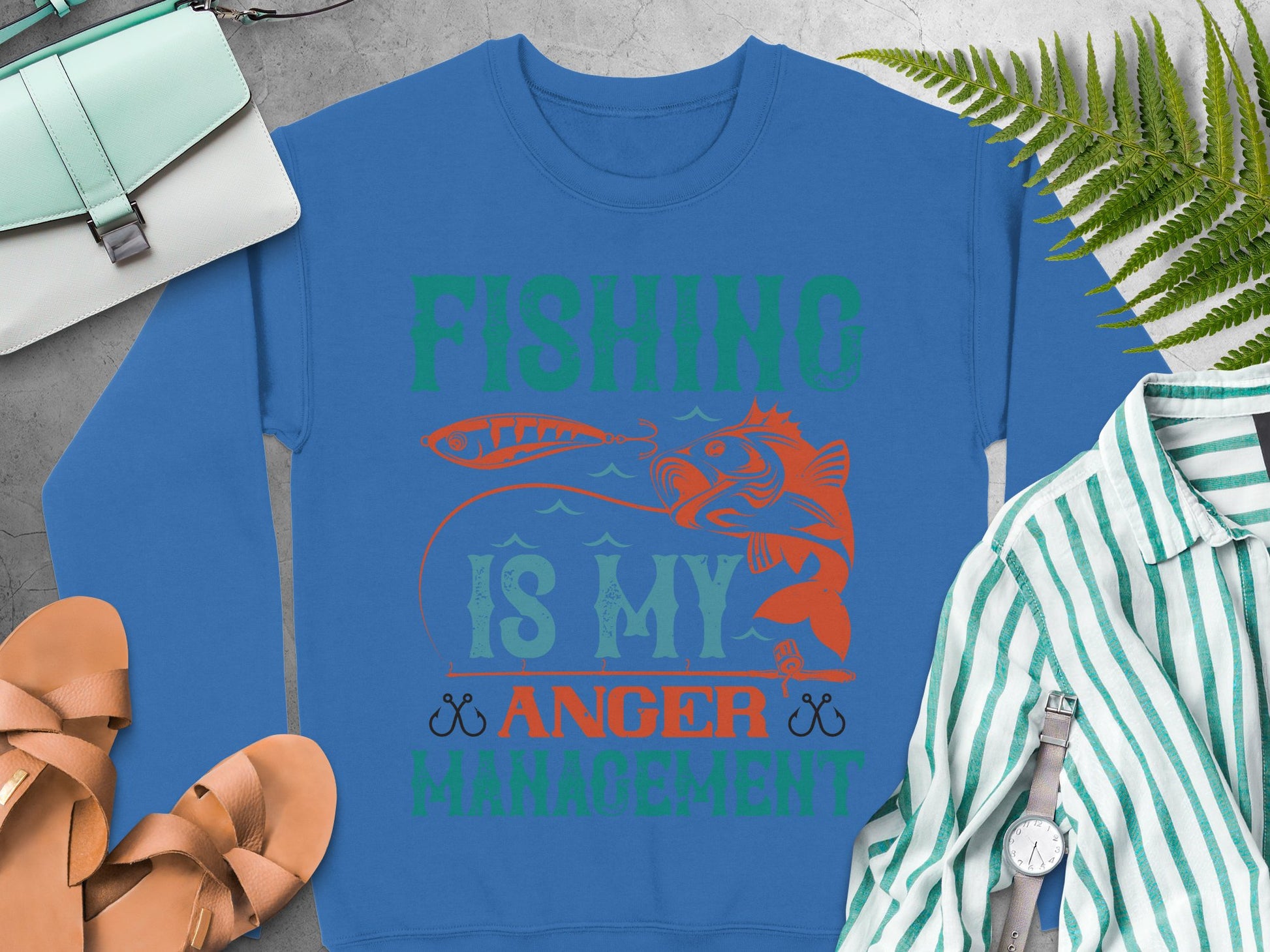 Experience Garment Graphics blue sweatshirt with the Fishing is my Anger Management design in vibrant green and orange. It features a fish and crossed hooks illustration, styled with sandals, a bag, striped shirt, watch, and fern leaves on a gray surface—perfect for fishing enthusiasts.