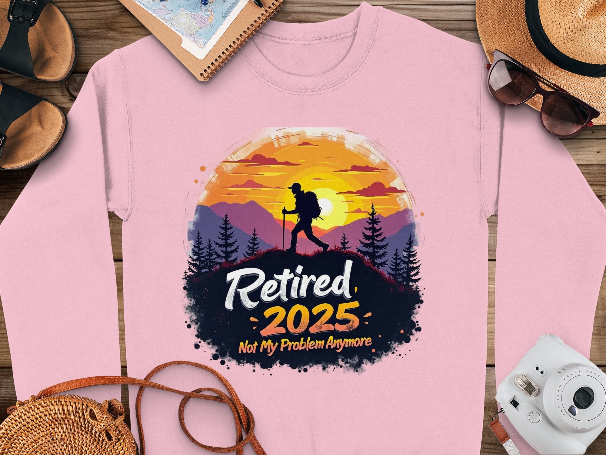Garment Graphics offers a pink retirement sweatshirt with a hiker design, featuring a person against a mountainous sunset and the text Retired 2025 Not My Problem Anymore. Perfect for adventurers, it pairs well with a straw hat, sunglasses, camera, brown shoes, and notebook. Unisex sizing available.