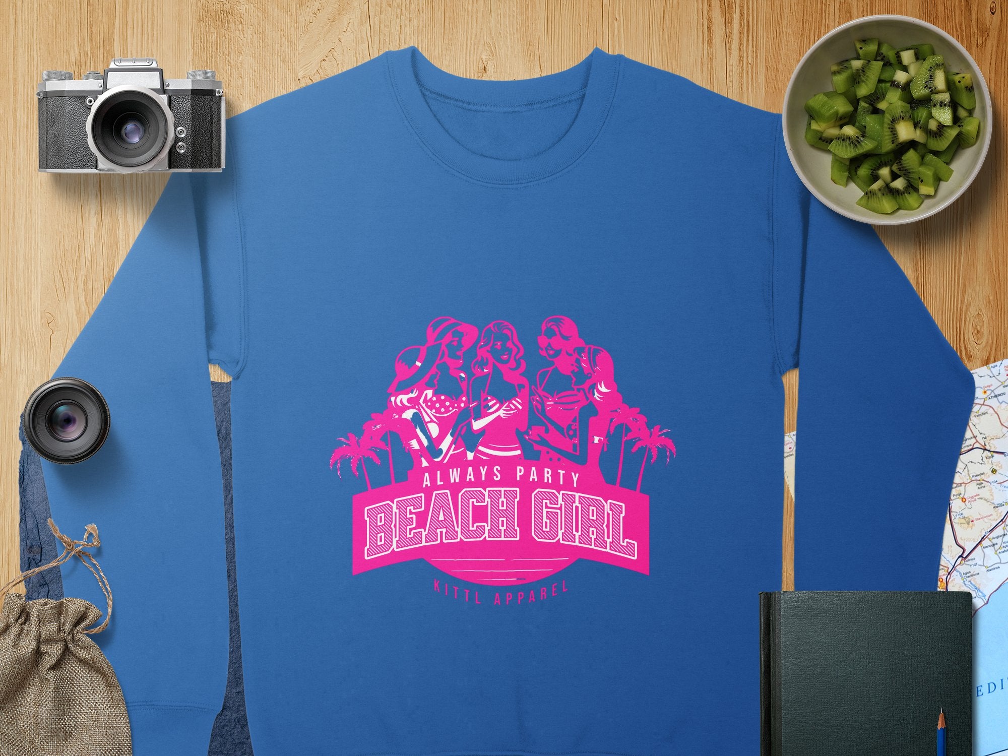 A blue sweatshirt from Garment Graphics features a pink graphic of three women and the phrases Always Party Beach Girl and Kittl Apparel. Surrounded by a camera, lens, sliced kiwi, notebook, bag, and map, this stylish piece captures the beachy charm perfectly.