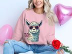 A woman in a whimsical Garment Graphics No Prob-Llama unisex sweatshirt, featuring a llama with sunglasses, holds a red rose while sitting next to pink heart-shaped balloons. Dressed in distressed jeans, she smiles at the camera, exuding charm and playfulness.