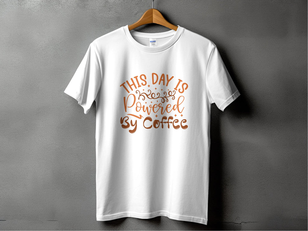 A white classic fit t-shirt from Garment Graphics hangs against a gray backdrop, displaying This Day is Powered by Coffee in bold brown cursive with decorative swirls—ideal for coffee lovers and positivity enthusiasts.