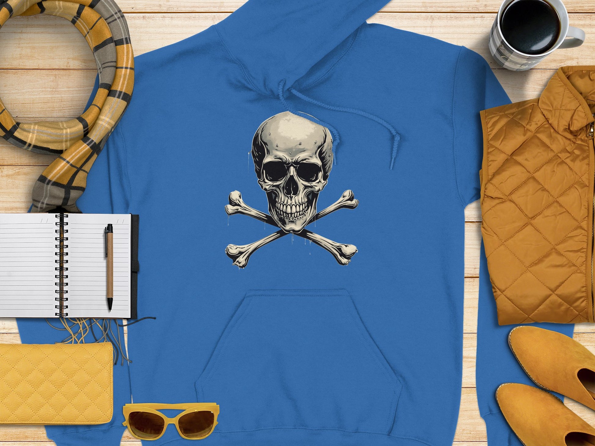 A blue Skull and Crossbones hoodie from Garment Graphics sits on a wooden surface, flanked by a yellow scarf, notebook, pen, yellow clutch, sunglasses, quilted jacket, shoes, and coffee mug—an edgy touch of comfort streetwear for any casual look.