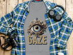 A Garment Graphics grey unisex t-shirt with an ornate eye and the word GAZE laid on wood, paired with a blue plaid shirt, headphones, camera, and coffee cup for a versatile style scene.