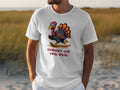 A person models a classic fit white Garment Graphics T-shirt featuring a funny cartoon turkey and the text TURKEY ON THE RUN against a coastal backdrop with tall grass and sandy terrain.