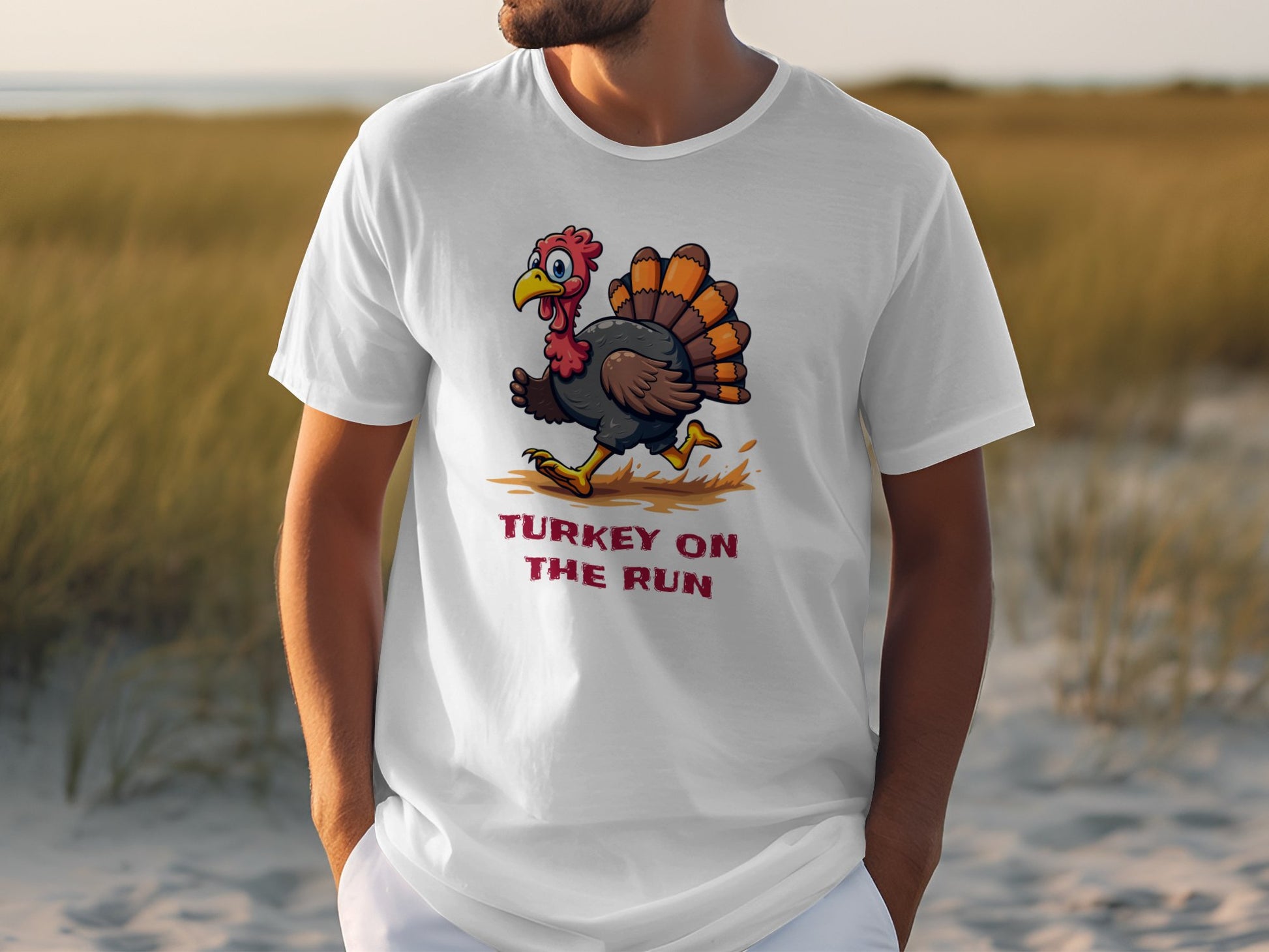 A person models a classic fit white Garment Graphics T-shirt featuring a funny cartoon turkey and the text TURKEY ON THE RUN against a coastal backdrop with tall grass and sandy terrain.