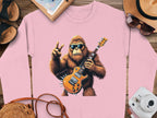 A pink sweatshirt from Garment Graphics showcases a gorilla wearing sunglasses, playing an electric guitar, and flashing a peace sign. Ideal for music lovers, it is styled on wood with a hat, sunglasses, a camera, sandals, and a notebook for a relaxed feel.