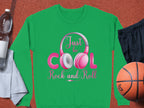 A green Garment Graphics sweatshirt features Just be Cool Rock and Roll in vibrant pink and white letters with Rock and Roll Pink Headphones. Ideal for music lovers, its paired on an orange surface with a water bottle, towel, black shorts, and a basketball—your stylish sporty-day statement.