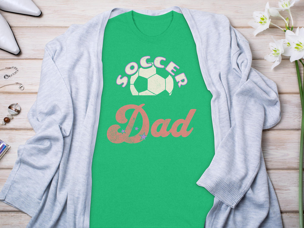 A green Garment Graphics T-shirt with Soccer Dad and a soccer ball design lies on a wooden surface, paired with a light gray cardigan. Nearby are white flowers, rings, and shoes that complete the charming display.