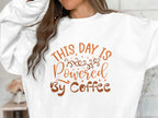 A person wears a Garment Graphics sweatshirt, featuring a lively design with This Day is Powered by Coffee in bright orange and brown, accented with swirls and stars. Only the torso and part of the arms are shown.