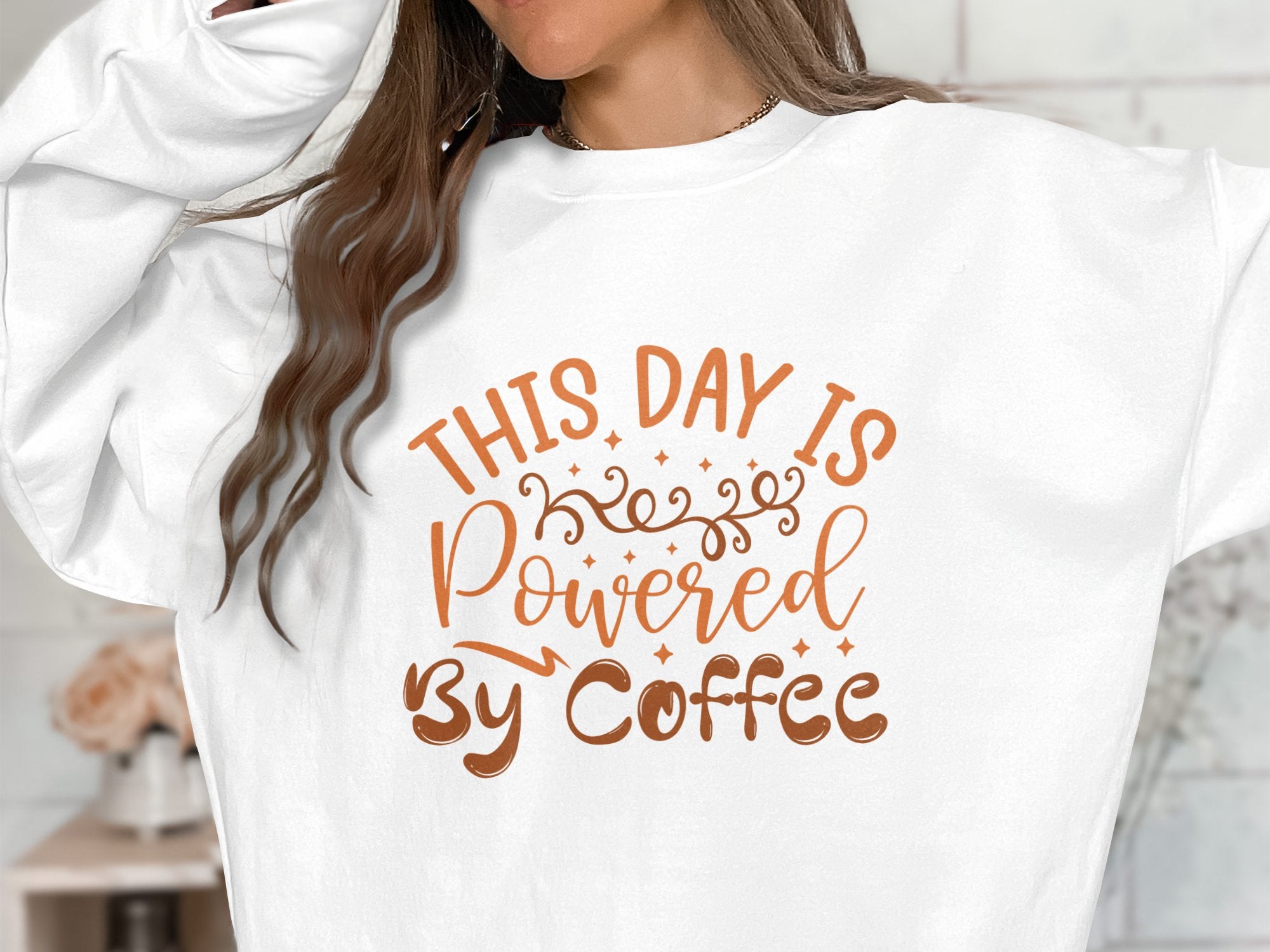 A person wears a Garment Graphics sweatshirt, featuring a lively design with This Day is Powered by Coffee in bright orange and brown, accented with swirls and stars. Only the torso and part of the arms are shown.