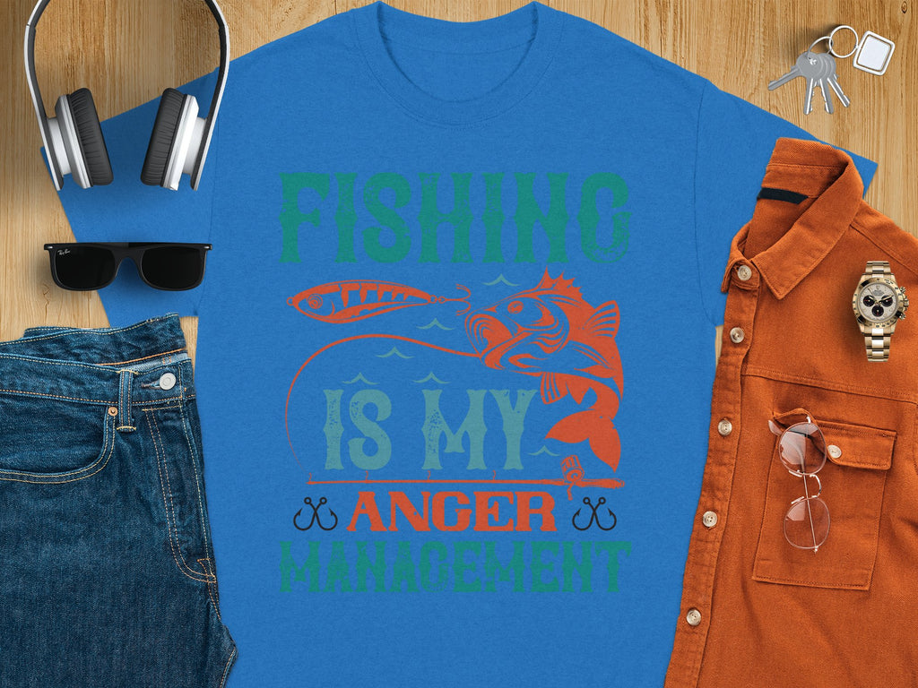 The Garment Graphics blue fishing T-shirt boldly states Fishing Is My Anger Management in vibrant green and orange, featuring a fish and lure. Paired with jeans, sunglasses, headphones, a watch, and a brown shirt on wood—ideal for any fishing enthusiasts casual day out.
