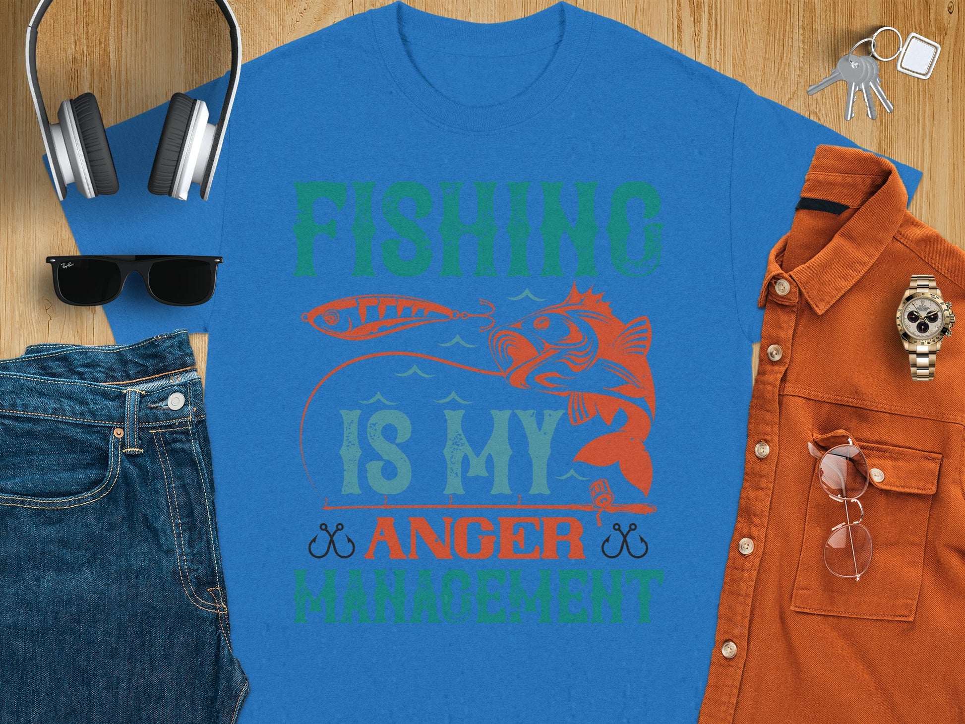 The Garment Graphics blue fishing T-shirt boldly states Fishing Is My Anger Management in vibrant green and orange, featuring a fish and lure. Paired with jeans, sunglasses, headphones, a watch, and a brown shirt on wood—ideal for any fishing enthusiasts casual day out.