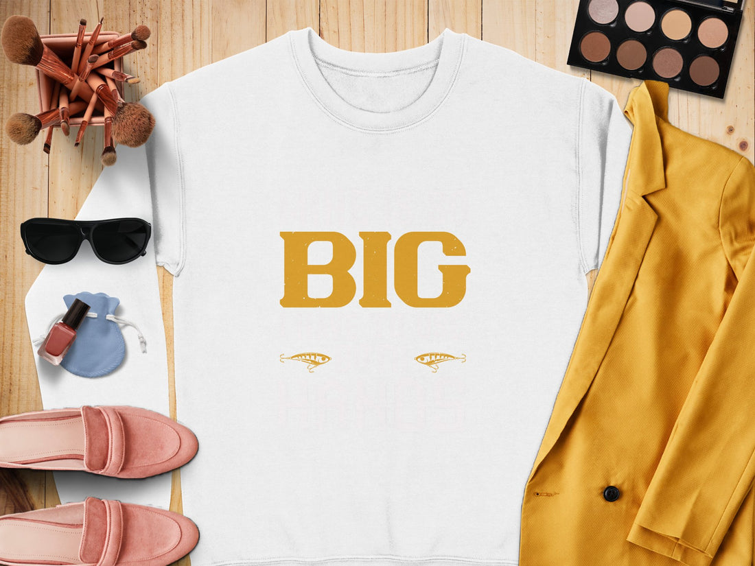 A Garment Graphics white shirt featuring BIG in large yellow letters and two eyes below rests on a wooden surface. Its styled with a yellow blazer, pink shoes, makeup brushes, sunglasses, an eyeshadow palette, a blue pouch, and nail polish—so big it takes both hands to hold!.