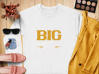 A Garment Graphics white shirt featuring BIG in large yellow letters and two eyes below rests on a wooden surface. Its styled with a yellow blazer, pink shoes, makeup brushes, sunglasses, an eyeshadow palette, a blue pouch, and nail polish—so big it takes both hands to hold!.