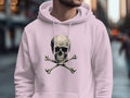 A fashion-forward individual wears a Garment Graphics light pink hoodie featuring a skull and crossbones design while standing on a bustling city street, with blurred skyscrapers and busy pedestrians enhancing the urban vibe.