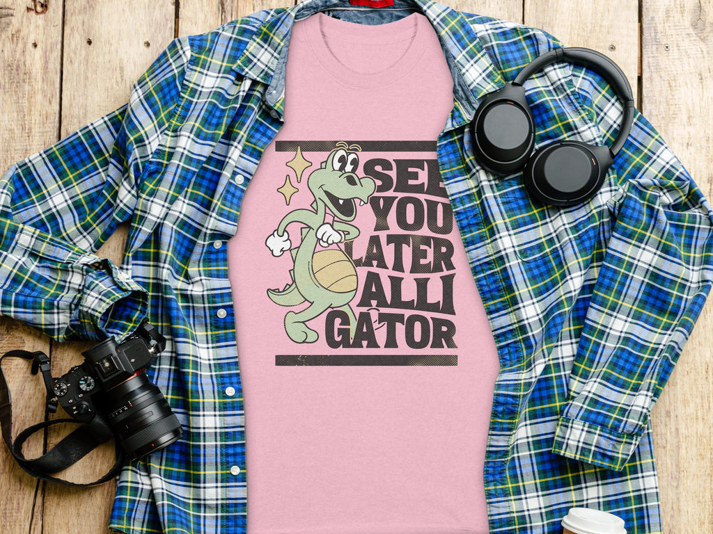 A Garment Graphics pink T-shirt with a cartoon alligator spinning a basketball and See You Later Alligator text is beneath a blue-green plaid shirt. Nearby, headphones, a camera, a pen, and a paper cup add to this casual ensemble on the wooden surface.