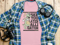 A Garment Graphics pink T-shirt with a cartoon alligator spinning a basketball and See You Later Alligator text is beneath a blue-green plaid shirt. Nearby, headphones, a camera, a pen, and a paper cup add to this casual ensemble on the wooden surface.