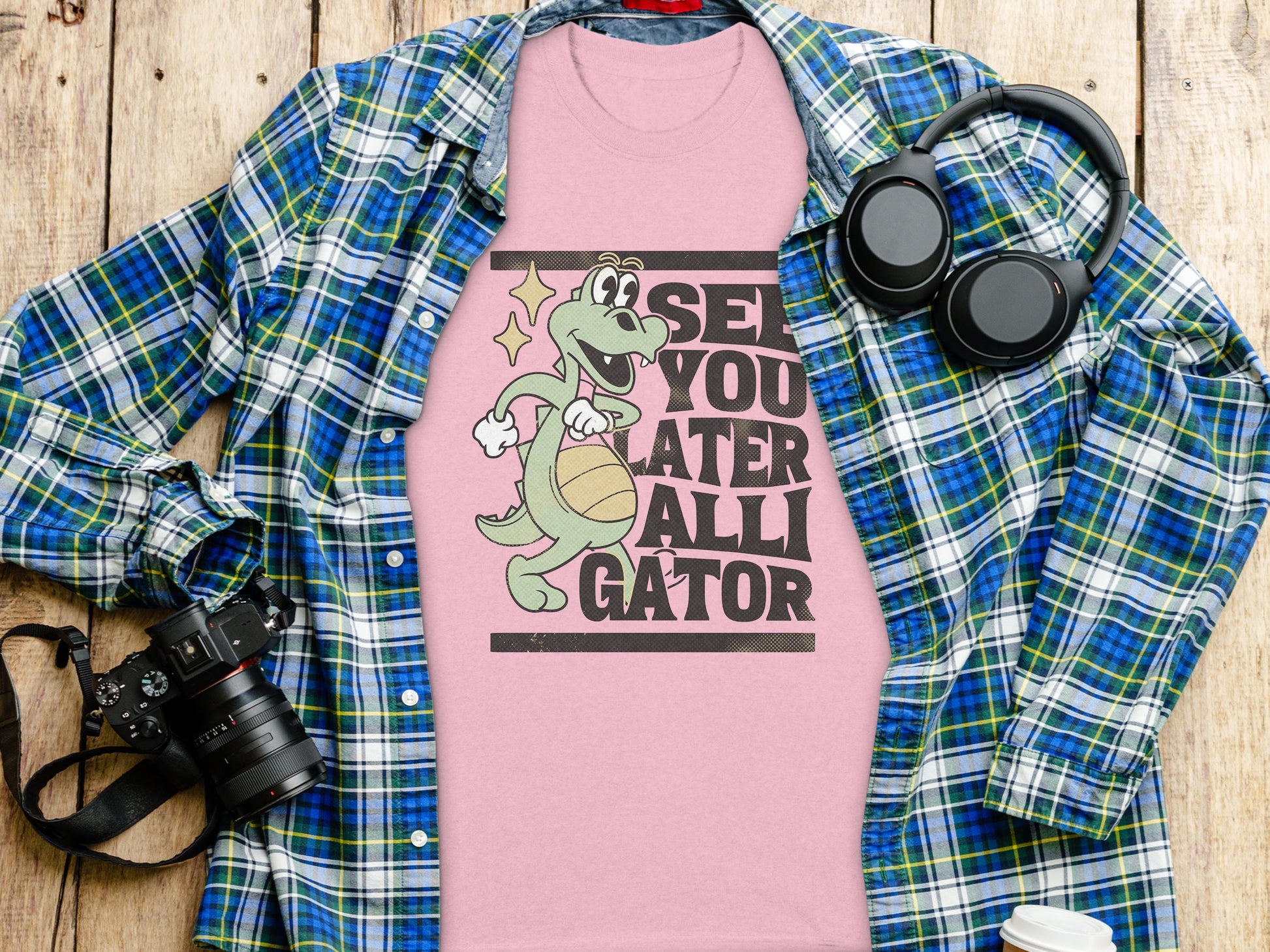 A Garment Graphics pink T-shirt with a cartoon alligator spinning a basketball and See You Later Alligator text is beneath a blue-green plaid shirt. Nearby, headphones, a camera, a pen, and a paper cup add to this casual ensemble on the wooden surface.