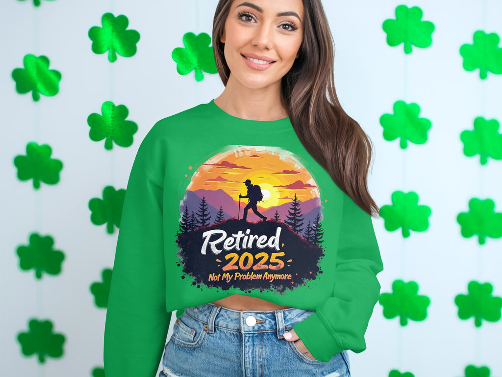A woman smiles in a Garment Graphics retirement sweatshirt, featuring a sunset hiker design with the text Retired 2025, Not My Problem Anymore. The unisex fit complements the green shamrocks in the background.