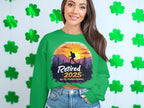 A woman smiles in a Garment Graphics retirement sweatshirt, featuring a sunset hiker design with the text Retired 2025, Not My Problem Anymore. The unisex fit complements the green shamrocks in the background.