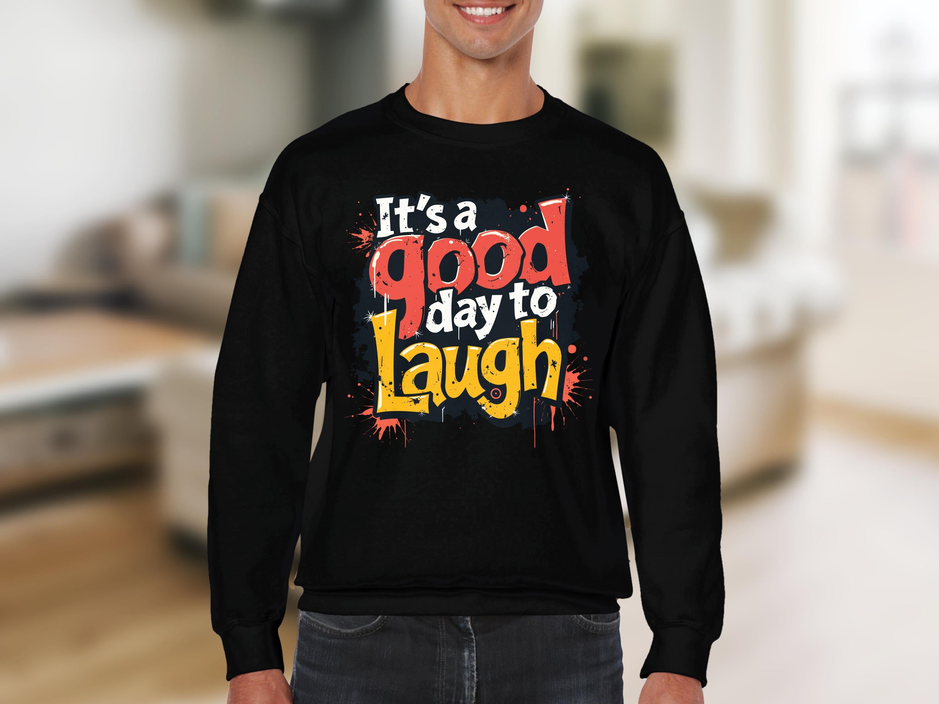 A person is sporting a black sweatshirt by Garment Graphics, adorned with vibrant graphics and the cheerful message, Its a Good Day to Laugh. The unisex fit ensures universal comfort. A softly blurred indoor setting in the background enhances its playful design.
