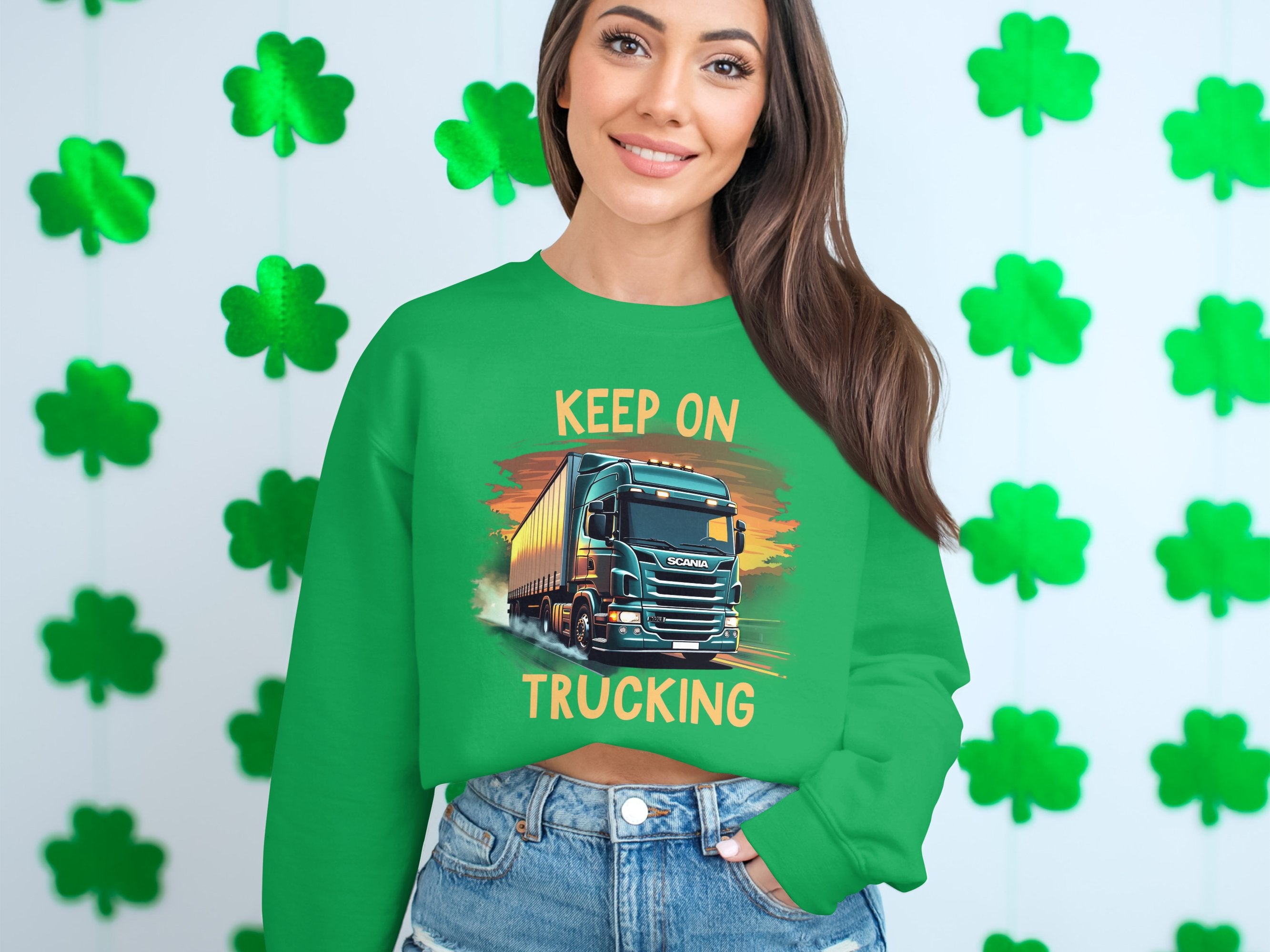 Against a clover-patterned wall, someone models Garment Graphics Graphic Sweatshirt for truck lovers, showcasing a bold truck design and KEEP ON TRUCKING text. This laid-back look is complete with long brown hair and denim shorts.